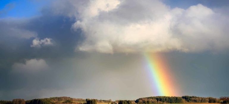 A rainbow emerging from a cloud over the countryside sky.
