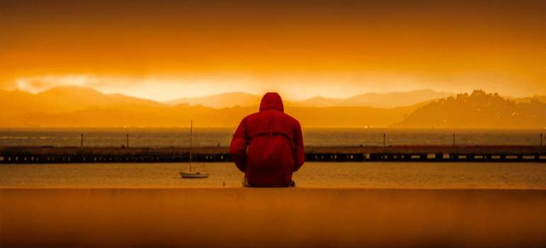 A hooded person sitting in front of a body of water.