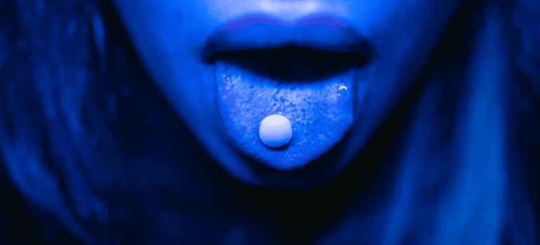 A close-up of a drug tablet in a person’s mouth under blue lighting.