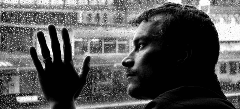 A black-and-white photo of a depressed man touching a window as it rains outside.