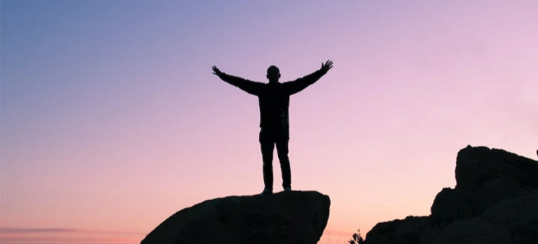 A silhouette of a man raising his hands as he stands on a rock.