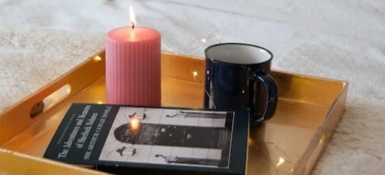 A bed table with a candle, a mug of coffee, and a book.