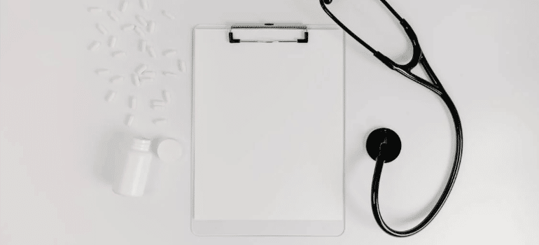 A white clipboard next to a stethoscope and a bottle of pills on a white surface.