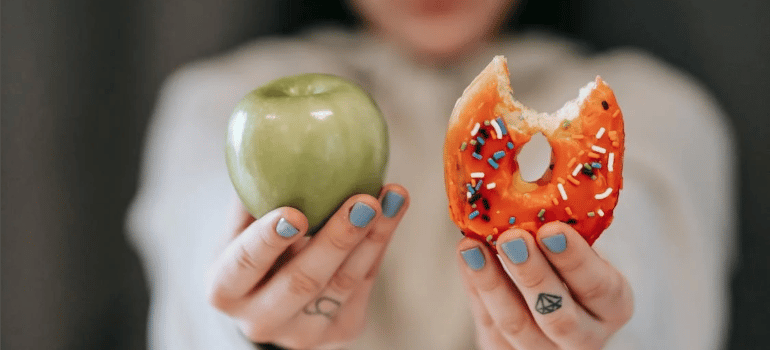 A close-up of a woman presenting an apple and a bitten doughnut.