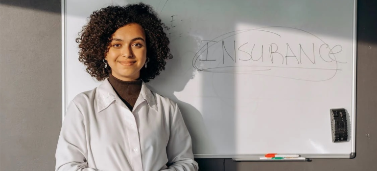 A smiling woman in a white coat next to a whiteboard that reads “insurance”.