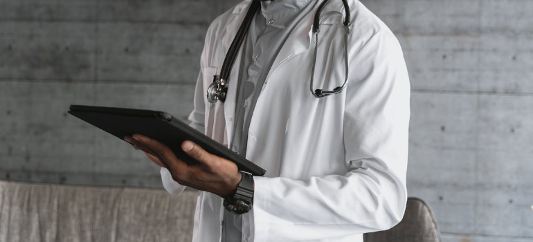 A doctor in a lab coat with a stethoscope around his neck holding a black tablet.