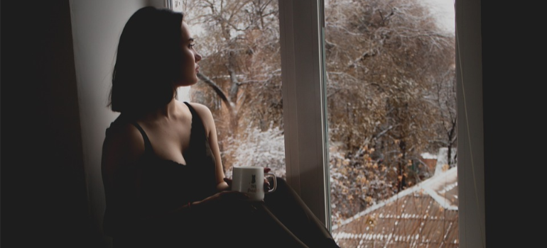 A close-up of a depressed young woman sitting by the window with a cup in hand before joining alcohol rehab center Buckhannon WV