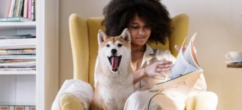 A woman in a yellow armchair, reading next to a happy dog.