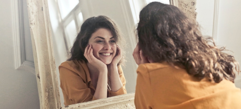 smiling woman looking at herself in the mirror, showing that addressing self-image issues is one of the unique needs women face in recovery