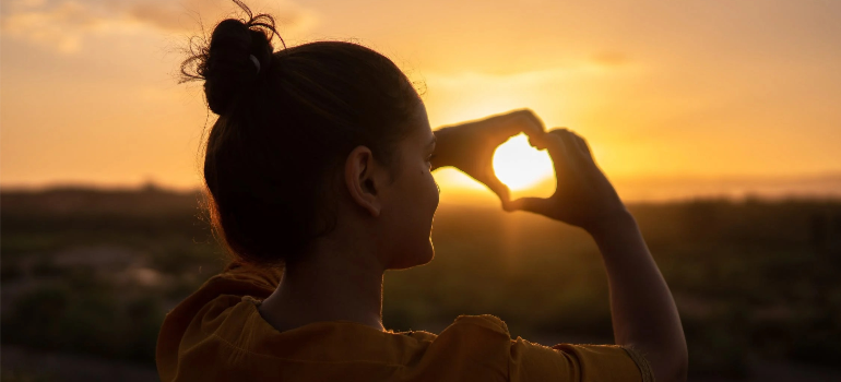 A woman doing a heart shape with her hands toward the sun.