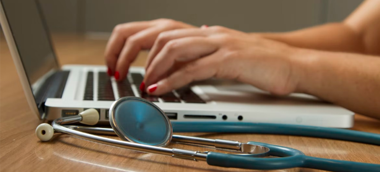 A close-up of a person using a laptop next to a stethoscope, symbolizing technology-assisted interventions for substance abuse.