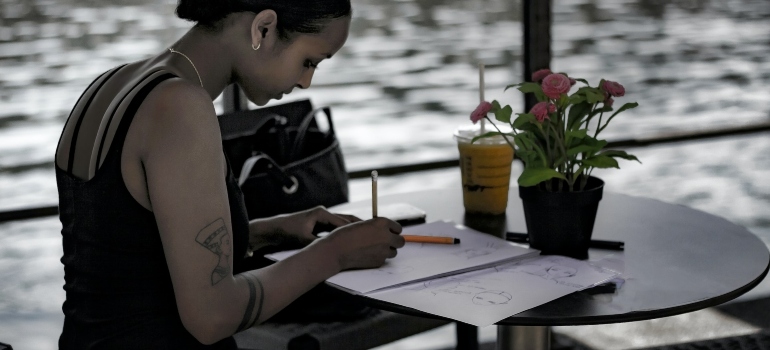 Person sitting at a table next to a body of water and writing into a journal, representing the power of positive thinking in addiction recovery