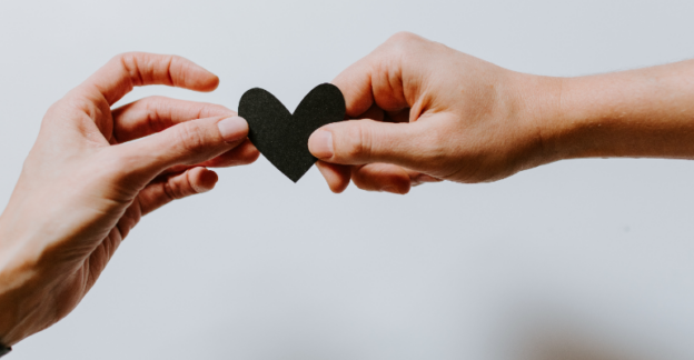 Two peoples hands holding together a small black paper heart.