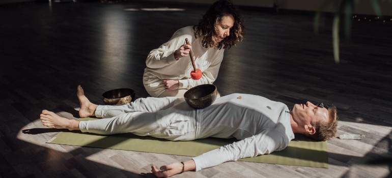 A man laying on a yoga mat while a woman is performing sound bath therapy