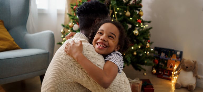 dad and daughter hugging each other with a christmas tree behind them representing the reasons why Holidays Are Difficult for People With Addictions