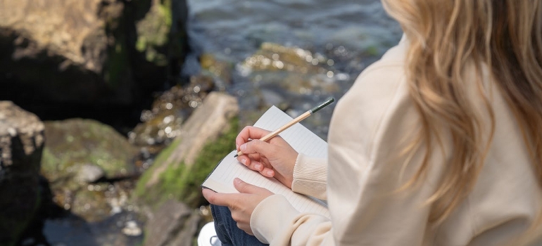 A woman is sitting by the water and writing a journal.