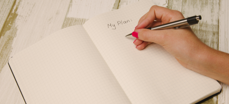 A person with red nails writes in their journal the words my plan