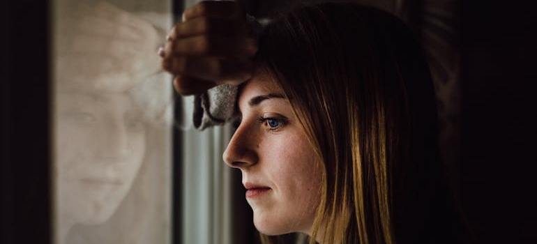 Woman thinking about whether she should move in with her parents after rehab and looking through the window.