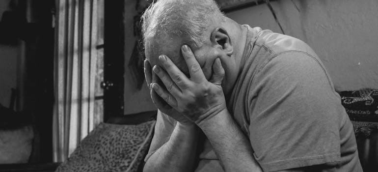 An older man sitting on the couch and holding his face in destress.