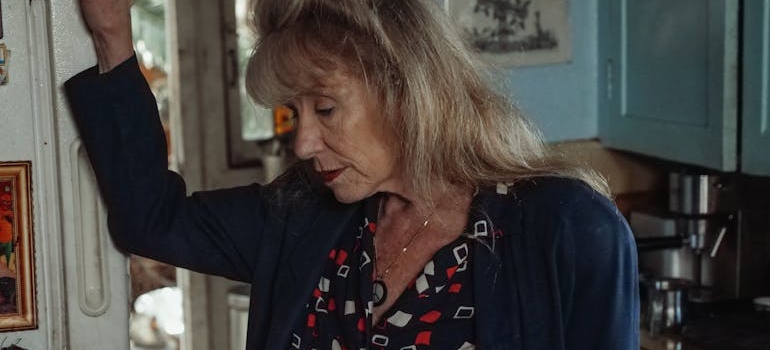 An older woman holding a door frame and looking down with a sad expression in her face.