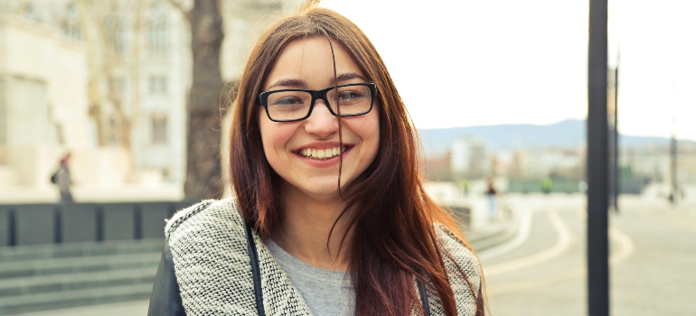 Smiling woman in gray jacket is standing near brown tree, successfully overcome addiction among Gen Z.