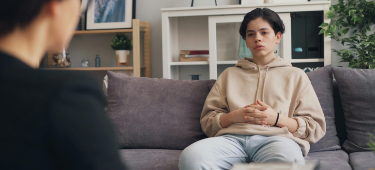 A boy in the beige sweatshirt is sitting on the couch and talking to a psychologist about how to educate kids on the dangers of alcohol.