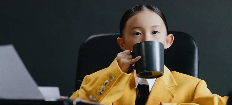 A little girl in the yellow jacket is drinking from a black cup.