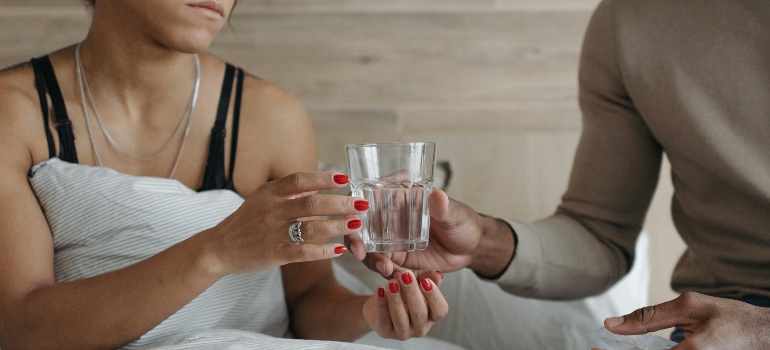 A man gives his wife a glass of water while sitting on the bed and talking about Restoril and melatonin.