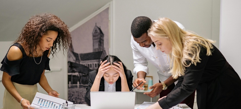 A woman sitting in front of a laptop and holding her head, with her colleagues standing next to her.