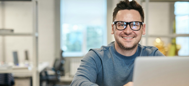 A man in a blue sweater wearing glasses and sitting in front of a laptop.