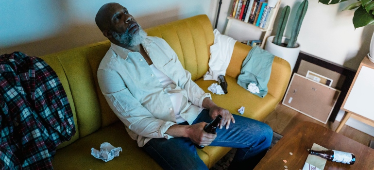 A depressed man is sitting on the couch and holding a bottle of alcohol.